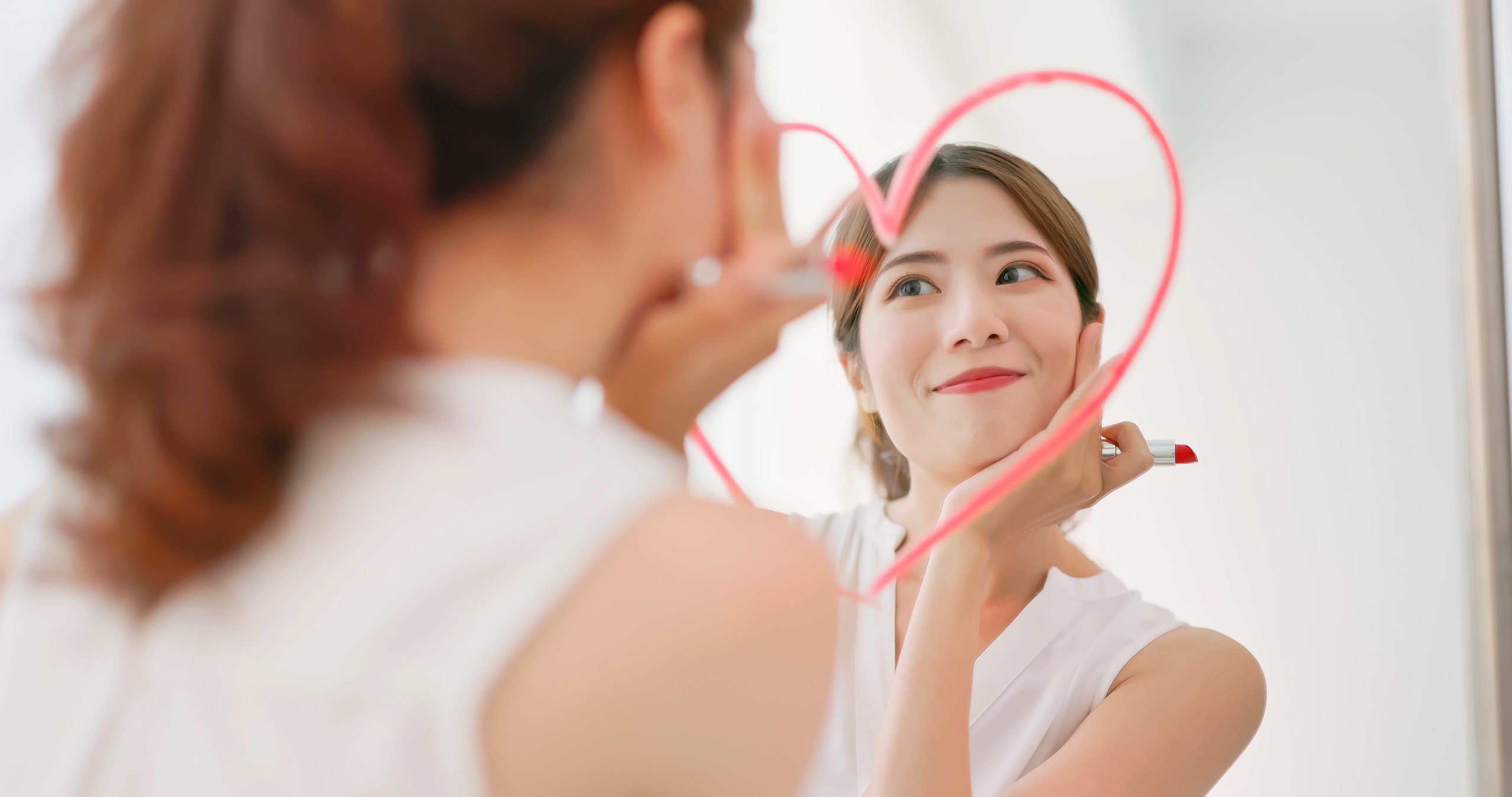 asiatische Frau mit Brunette-Ponytail zieht eine Herzform, benutzt Lippenstift auf dem Spiegel und betrachtet sich selbstbewusst Selbstliebe